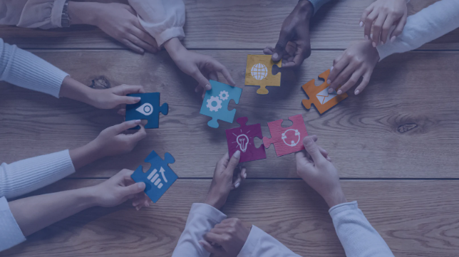 Obraz pokazuje osiem rąk trzymających kolorowe puzzle z ikonami symbolizującymi różne obszary pracy zespołowej, jak analiza, innowacje, technologia czy globalna współpraca. To metafora wspólnego działania i łączenia kompetencji.